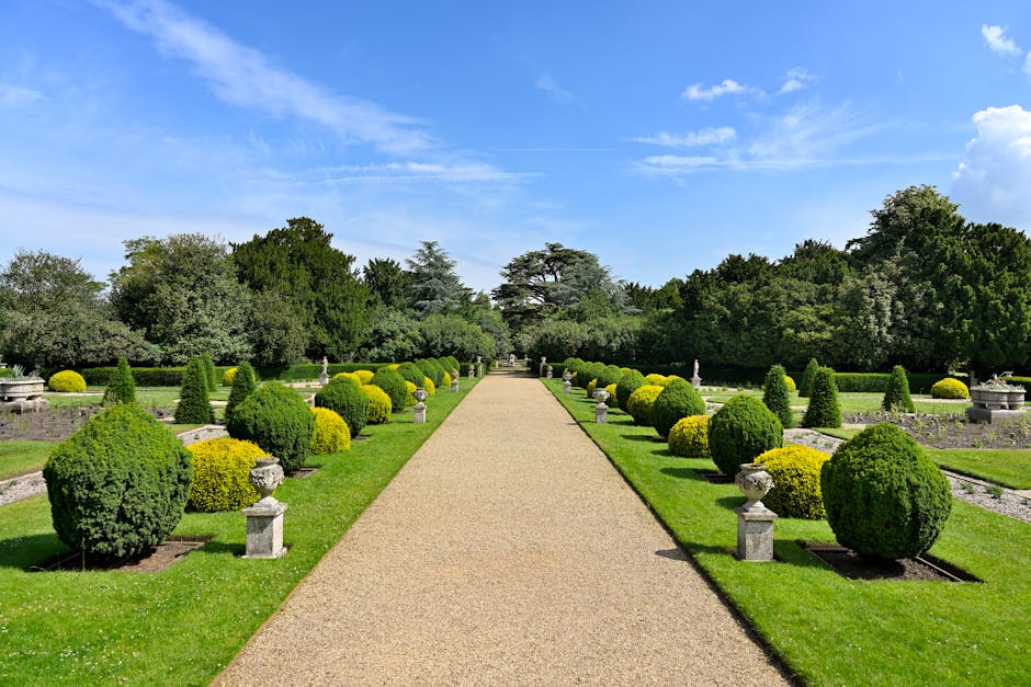 A lush garden scene with a well-maintained, green lawn bordered by a variety of flowering plants and shrubs. Tall trees with twisting branches and dense foliage provide shade, while a vibrant array of
